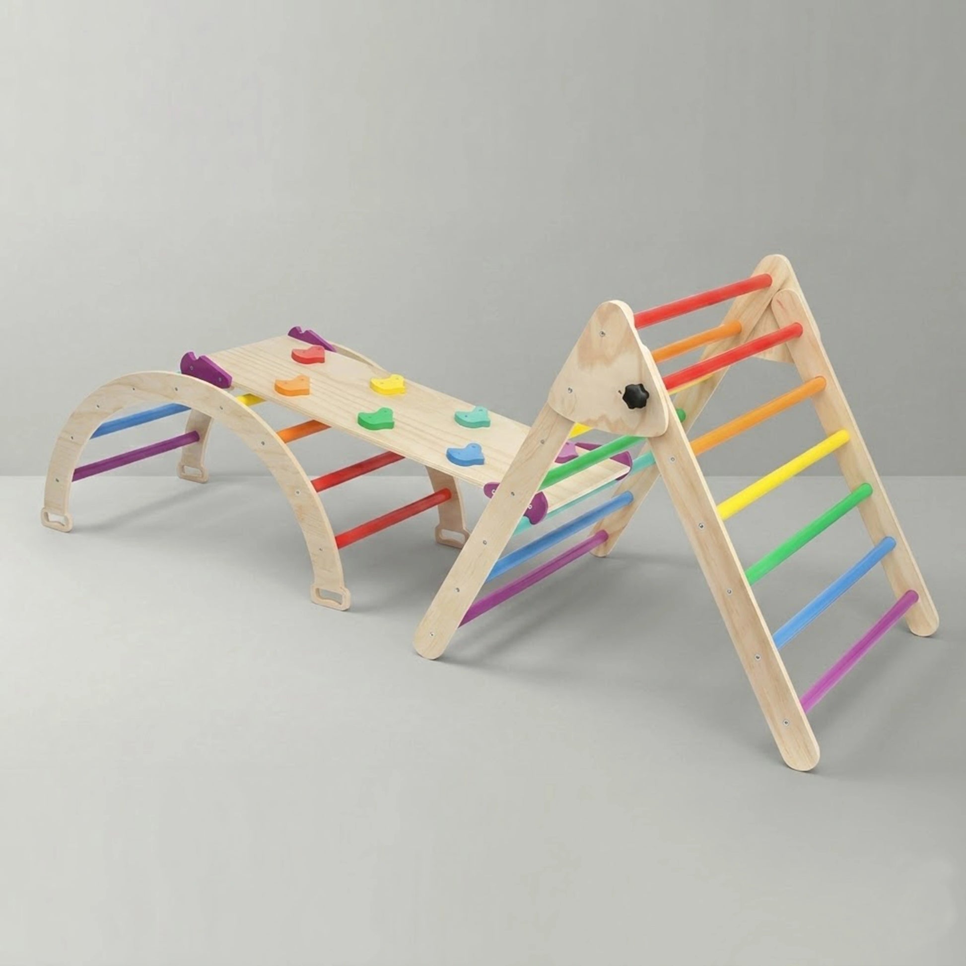 A studio shot of a 3-piece wooden Montessori climbing set against a clean grey background. The indoor playground features a climbing arch and a triangle climber, both with bright rainbow-colored rungs, securely connected by a reversible wooden ramp with colorful rock climbing holds.