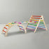 A clean studio shot of a 3-piece children's wooden Montessori climbing set against a light grey background. It clearly displays the climbing arch on the left and the Pikler triangle on the right, both with bright rainbow rungs, seamlessly connected by a wooden rock climbing ramp.