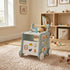 A colorful wooden 6-in-1 toddler walker and play kitchen activity center on a white studio background. Features include a push handle with colorful rings, a toy storage box, a teaching clock, shape matching, and a stove with a sink. Suitable for ages 12+ months.