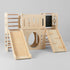 A studio shot of a wooden indoor castle slide for kids against a grey background. The angle highlights the long, smooth slide on the left, the central play tunnel, and the sturdy climbing ramp on the right side.