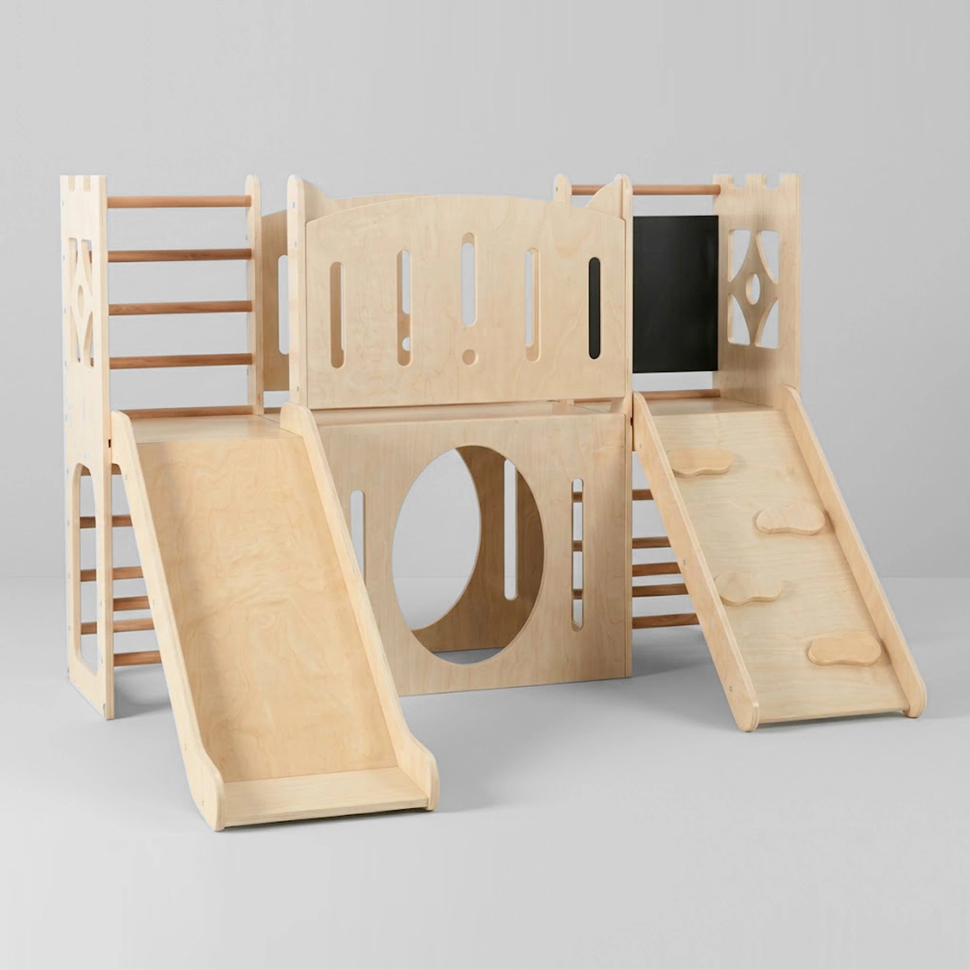 A studio shot of a wooden indoor castle slide for kids against a grey background. The angle highlights the long, smooth slide on the left, the central play tunnel, and the sturdy climbing ramp on the right side.
