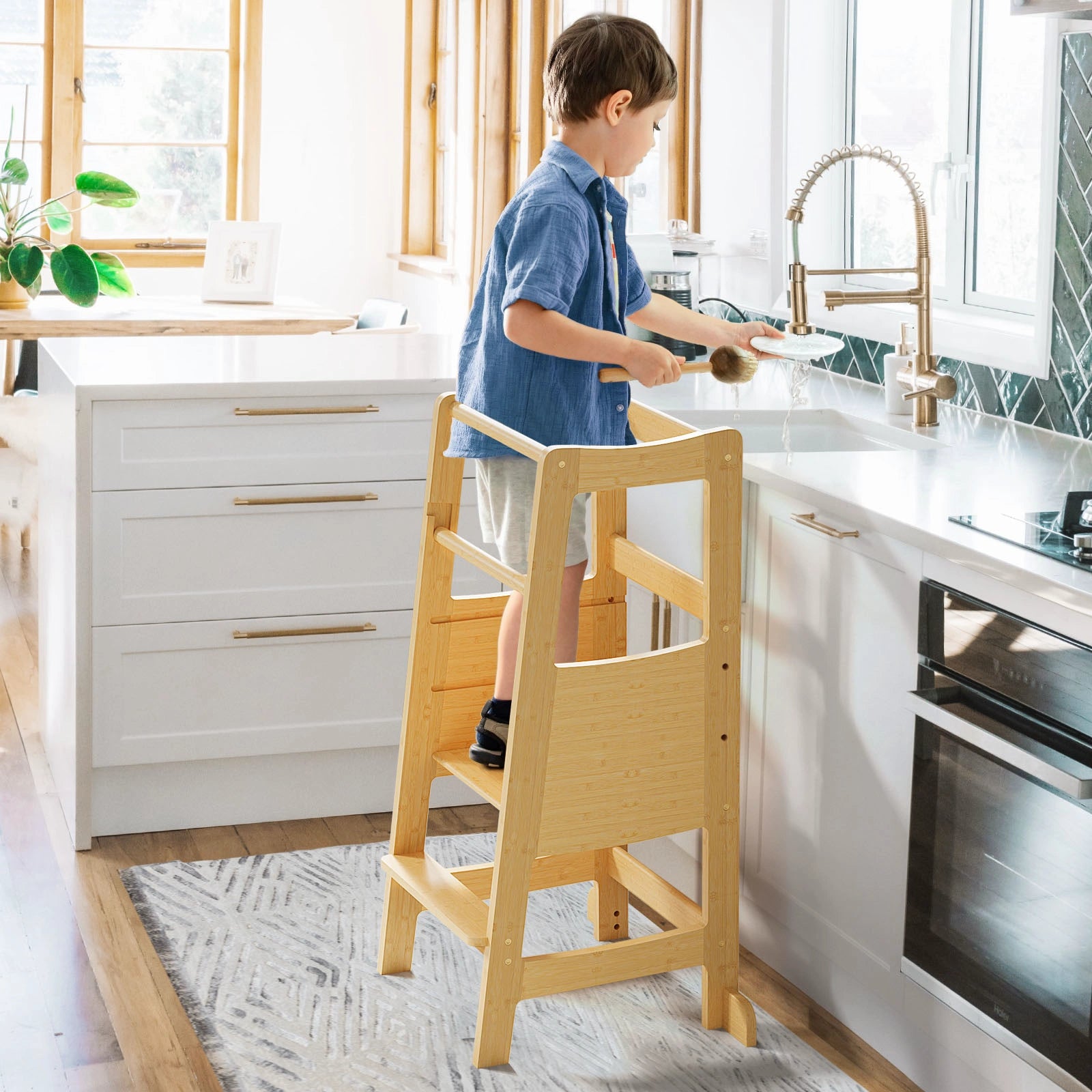 A young boy stands safely on a natural bamboo toddler learning tower, helping to wash dishes at a modern white kitchen sink.