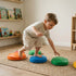A barefoot toddler boy in a striped shirt is balancing on a colorful blue stepping stone while reaching down with one hand to touch a green textured stone. He has one foot on the blue stone and one foot on an orange stone behind him. The stones are scattered on a woven rug in a sunny playroom with wooden bookshelves, demonstrating active play and sensory exploration.