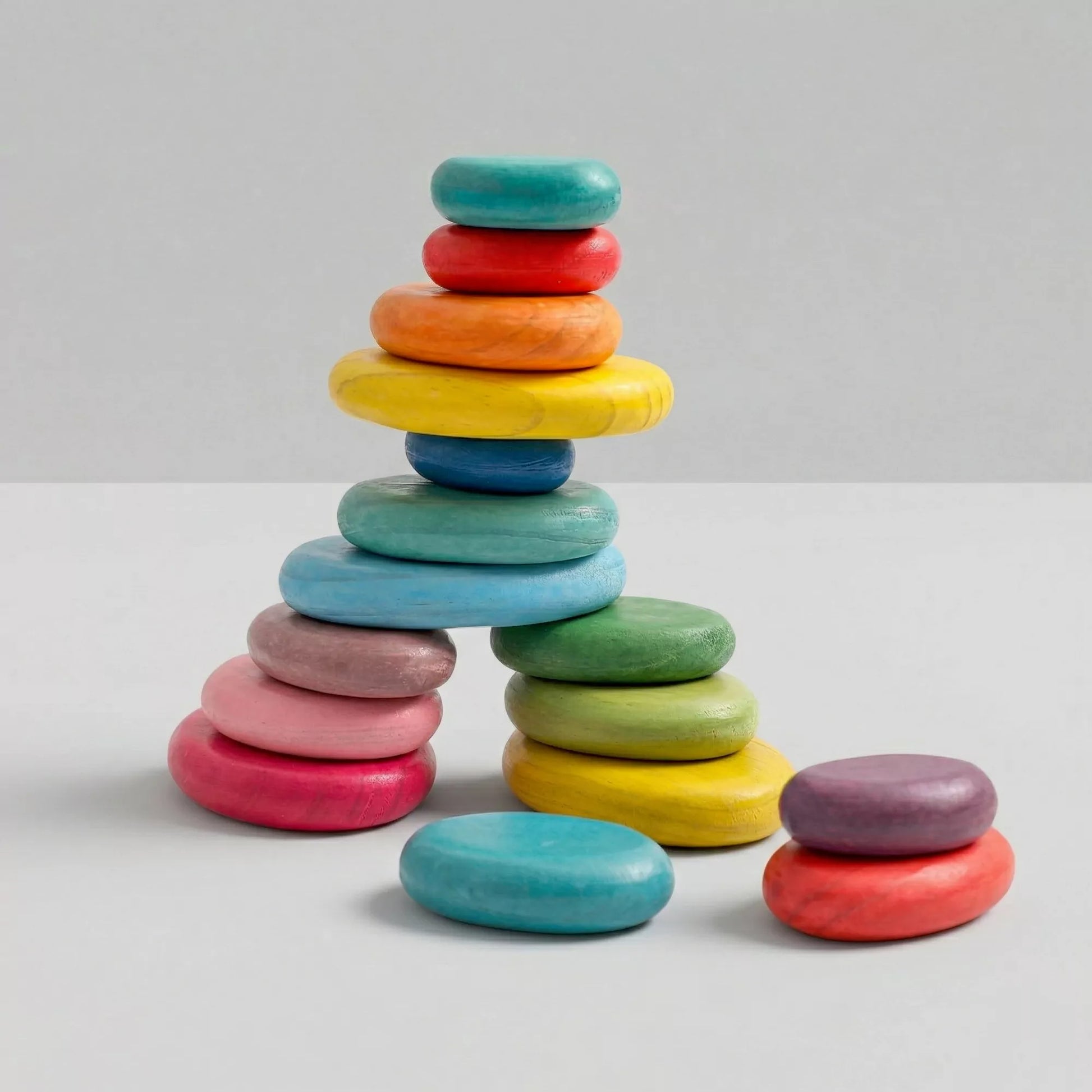 A collection of smooth, colorful wooden balancing stones. Several stones are stacked to form an arch-like structure on a plain white surface, with a few loose stones resting beside it.