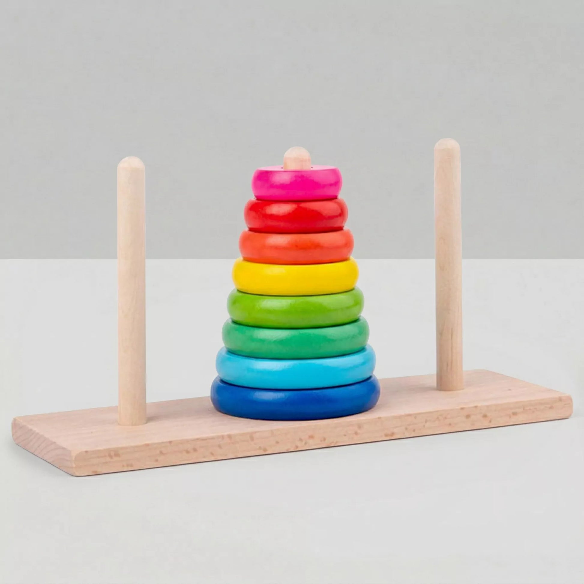 A wooden educational stacking toy with a rectangular base and three pegs, displayed against a plain white background. The center peg holds a stack of brightly colored wooden rings arranged by size to form a pyramid.