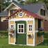 A scenic outdoor photograph showing a beige and gray wooden cottage-style convertible playhouse and chicken coop sitting on a grassy lawn in a sunny backyard. It features a main double door with glass panes, four front windows with white trim and attached planter boxes filled with flowers, and a gray roof.