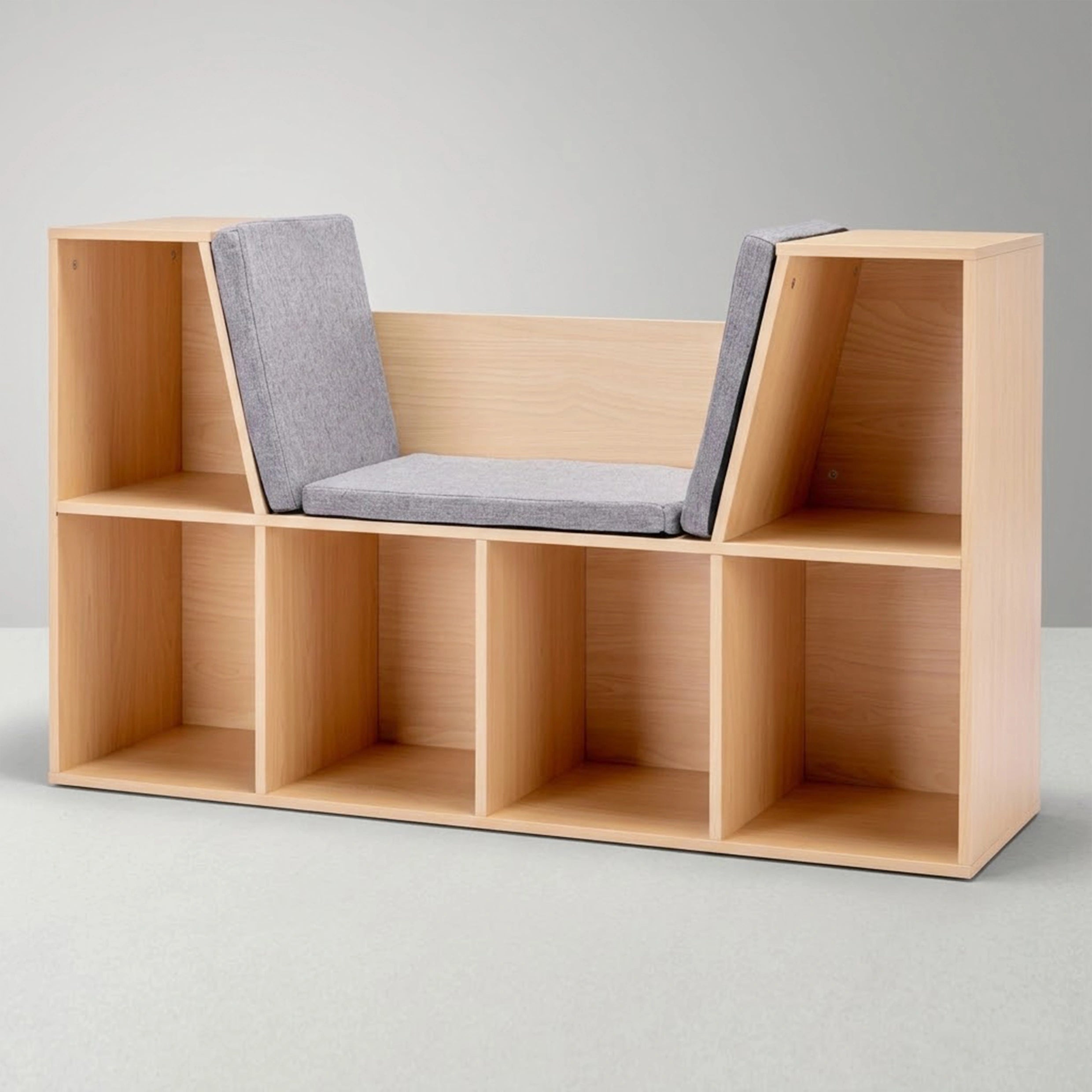 A studio shot of an empty, light wood kids' bookcase against a clean grey background. The furniture piece features a central built-in reading nook with grey cushioned seating, surrounded by six open bottom cubbies and two upper side shelves for storage.