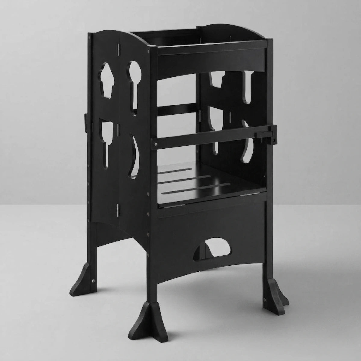 A black wooden folding toddler learning tower shown from a slight front angle on a light grey background, featuring safety bars and decorative kitchen utensil cutouts on the sides.