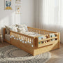 Cozy toddler bedroom decor featuring the Hicooo wooden Montessori floor bed. The bed has safety rails, an attached footboard bookshelf, and is decorated with string lights, sitting on a round rug in a sunlit room.