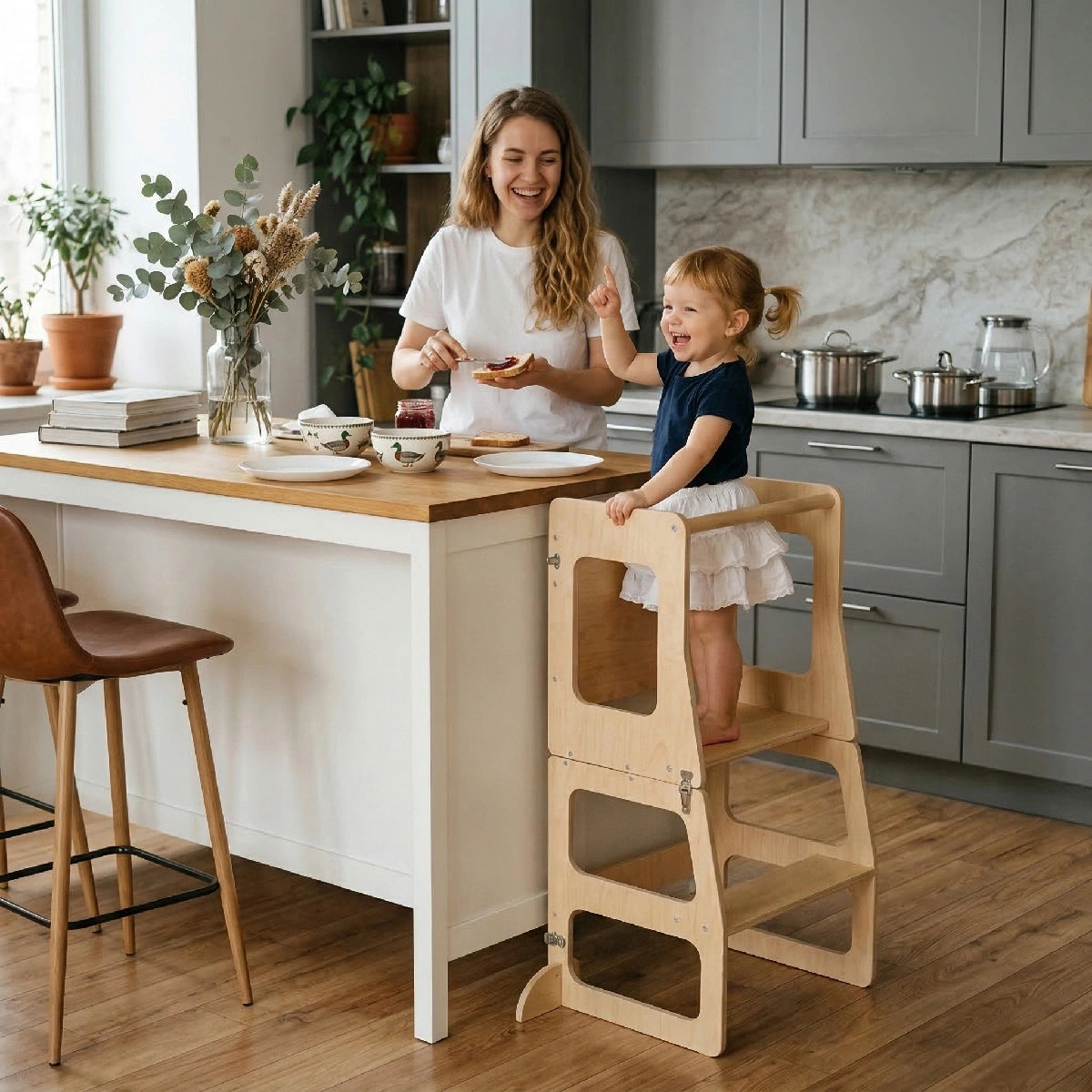 A laughing toddler stands safely in the Hicooo natural wood kitchen helper tower at a kitchen island, excitedly pointing while her mother prepares toast with jam nearby.