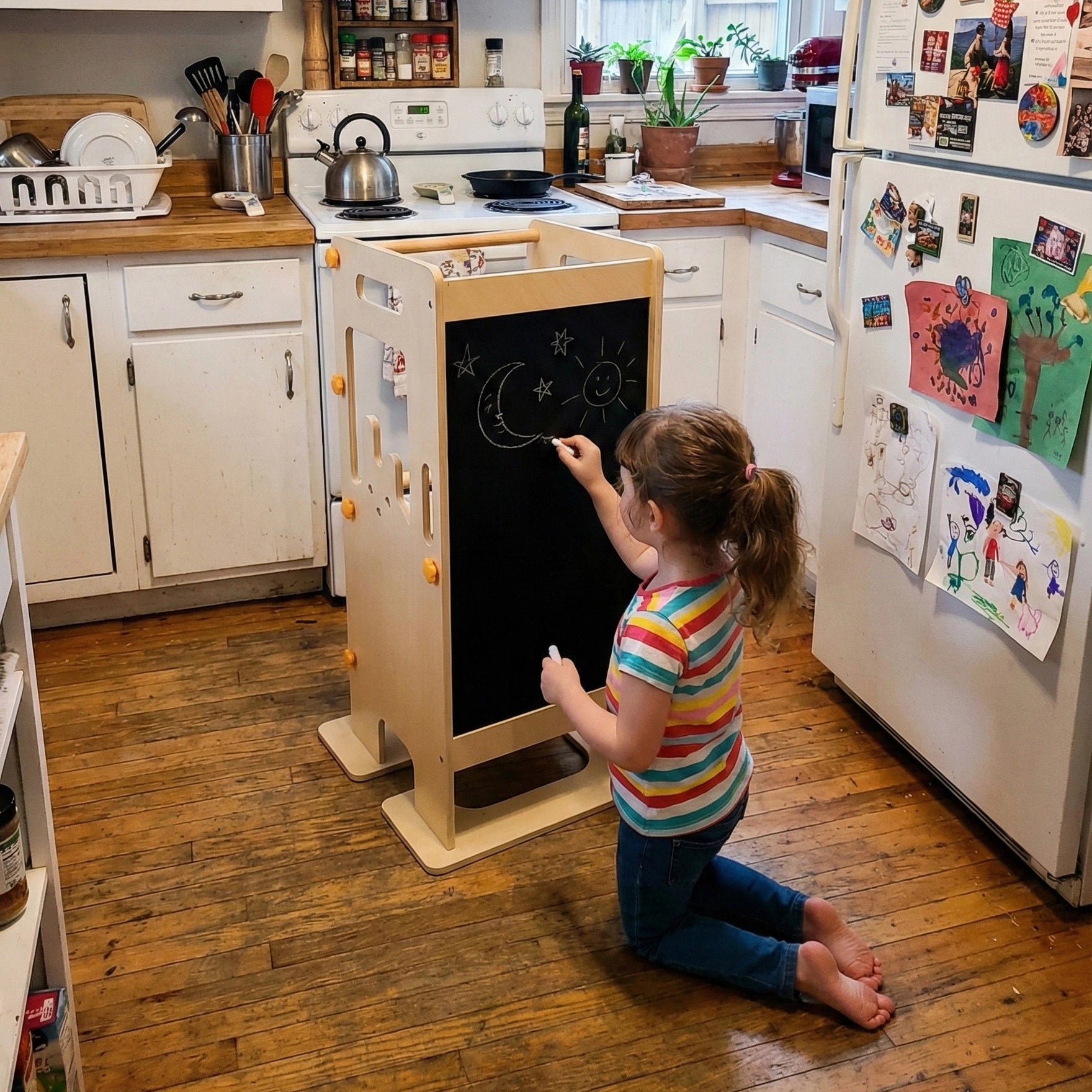 Little girl kneeling on the kitchen floor drawing a moon and stars on the built-in chalkboard back of the HICOOO wooden learning tower. Shows multi-functional design for creative play.