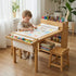 Lifestyle photo of a toddler boy in overalls drawing with crayons on the Hicooo wooden art table. The desk features a continuous paper roll for drawing, a top organizer tray filled with markers, and sturdy wooden stools.
