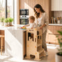 A smiling mother watches her toddler reach for vegetables on the kitchen island while standing safely in a HICOOO wooden rabbit learning tower. Perfect for Montessori-style kitchen bonding and healthy eating habits.