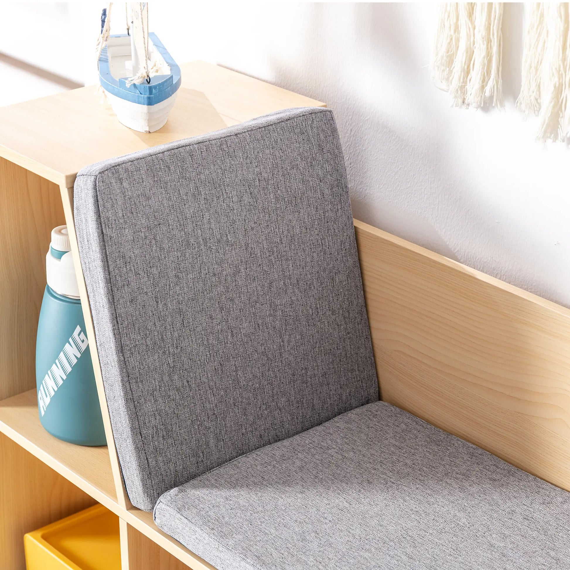 A close-up view of the built-in reading bench on a light wood kids' bookcase, focusing on the comfortable grey fabric cushions for the seat and angled backrest. A blue water bottle is conveniently stored in the adjacent open cubby, highlighting the practical and ergonomic design.