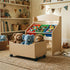 A cozy playroom setting featuring a light wood kids' toy storage organizer. The large bottom wooden toy box on casters is pulled out onto a woven rug, filled with plush animals including an elephant and a lion. Above, three colorful removable bins in red, green, and blue neatly organize building blocks, animal figures, and art supplies.
