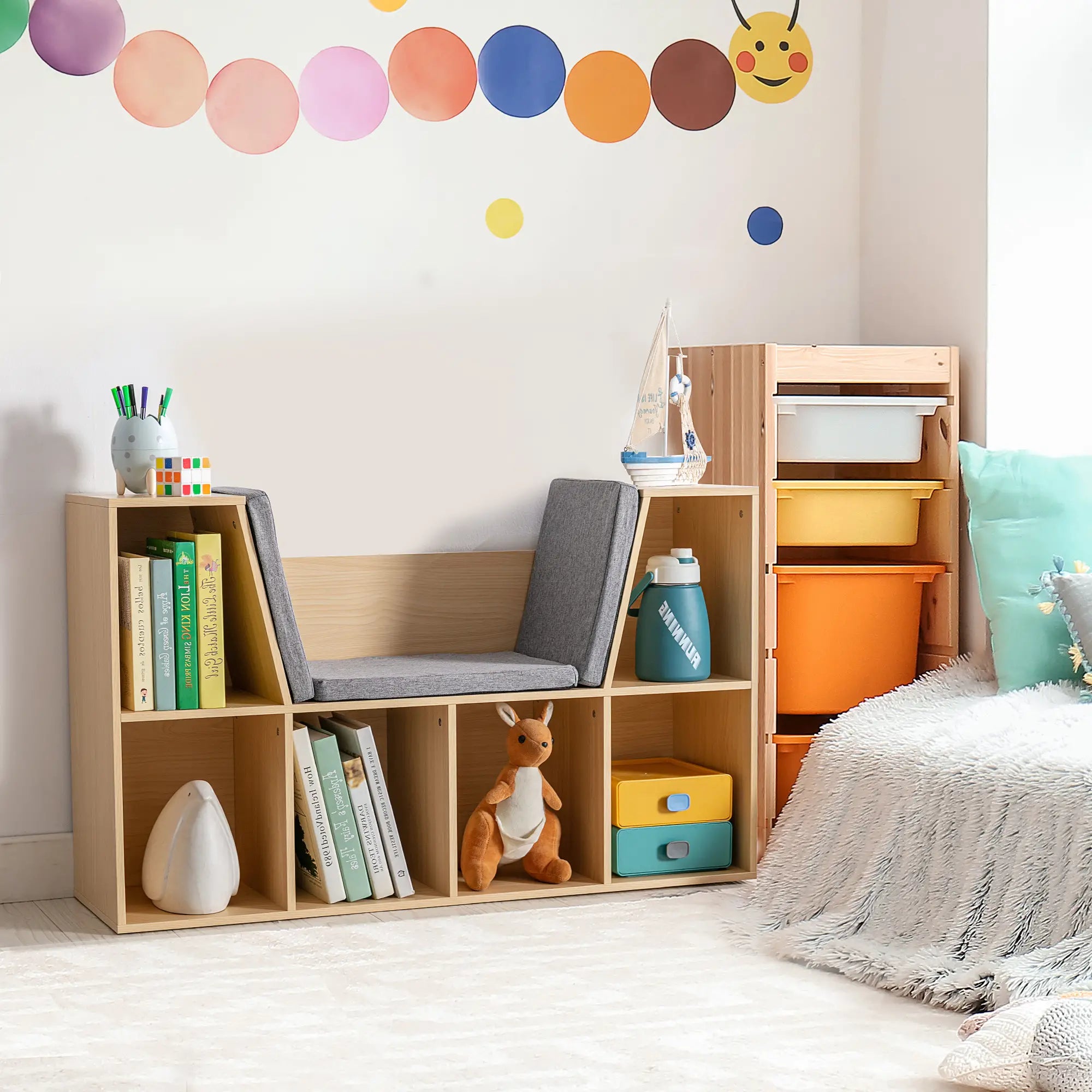 A beautifully styled light wood kids' bookshelf with a built-in grey reading bench in a vibrant playroom. The open cubbies are decorated with books, a kangaroo plushie, and small toy boxes, placed next to a colorful storage bin organizer.