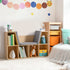 A beautifully styled light wood kids' bookshelf with a built-in grey reading bench in a vibrant playroom. The open cubbies are decorated with books, a kangaroo plushie, and small toy boxes, placed next to a colorful storage bin organizer.