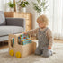 Toddler kneeling on a rug, selecting a picture book from a Hicooo wooden mobile book caddy with yellow wheels in a sunny living room.