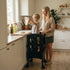 A smiling mother watches as her young child, standing in a black wooden toddler tower, excitedly stretches pizza dough at a bright kitchen counter near a window.