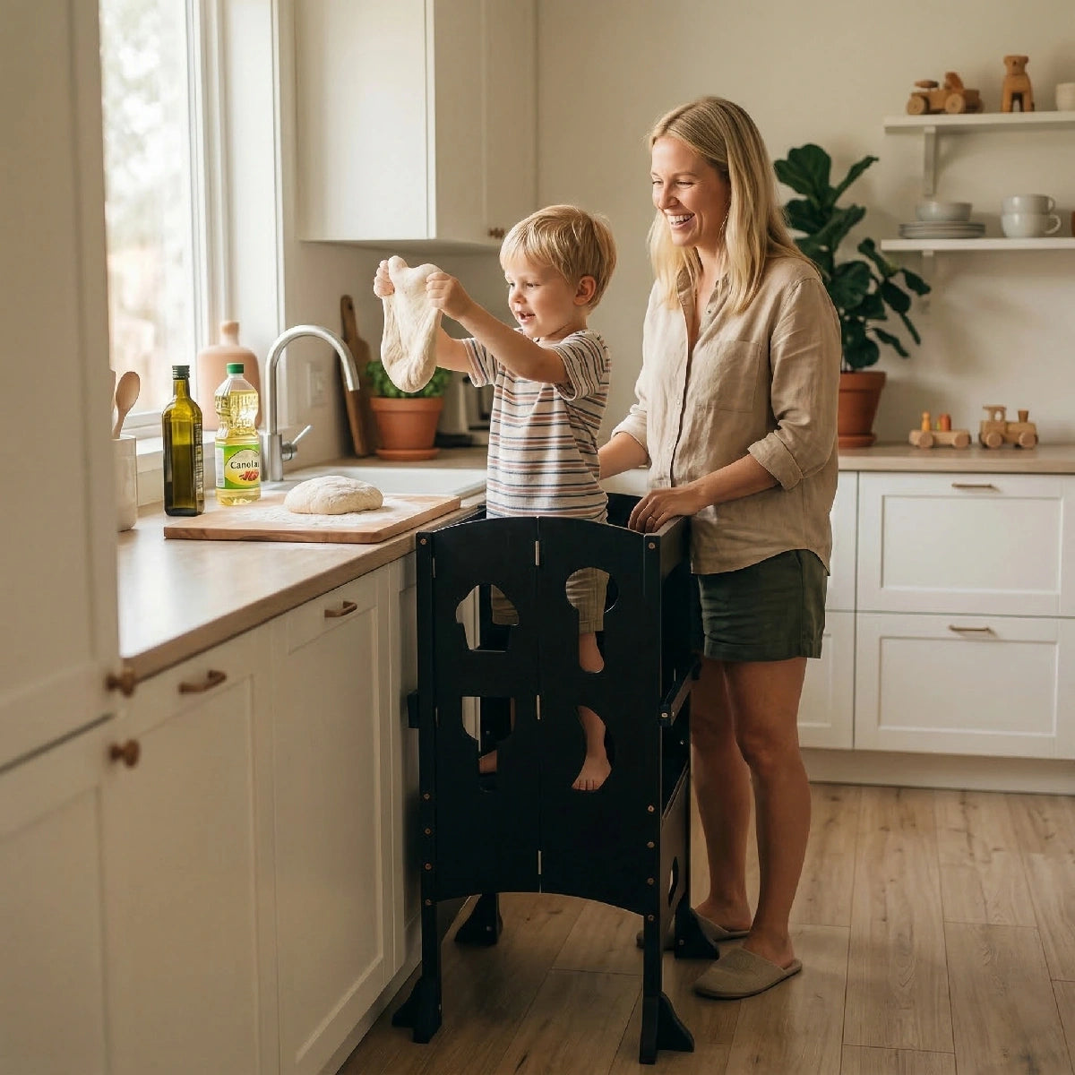 A smiling mother watches as her young child, standing in a black wooden toddler tower, excitedly stretches pizza dough at a bright kitchen counter near a window.