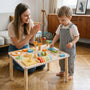 A mother claps and cheers for her toddler son who is standing at a hicooo wooden Montessori activity table. The child is holding a blue triangle block, engaging in shape sorting play.