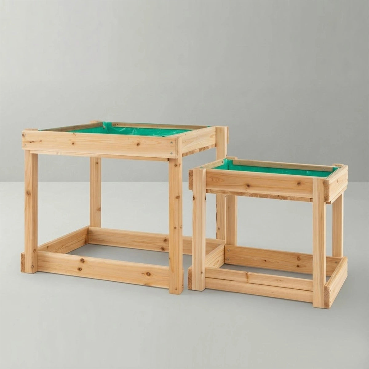 A clean studio shot of a two-piece nesting wooden sand and water sensory table set for kids. Crafted from light fir wood, it features one taller table and one shorter table designed to slide and fit neatly underneath for convenient, space-saving storage. Both table beds are lined with a green waterproof liner, ready for messy play against a plain light gray background.