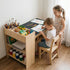A toddler boy sits at Hicooo wooden art desk drawing, while his older sister stands behind him smiling. The desk features a large side storage organizer filled with art supplies, highlighting a perfect shared workspace for siblings.