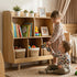 A heartwarming playroom scene featuring siblings playing in front of the Hicooo wooden wavy kids' bookshelf. A toddler girl watches her baby brother play with a toy car near the toy organizer filled with books and baskets.