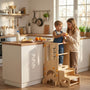 Older sister sharing an orange slice with her toddler brother who is standing safely at counter height in a Hicooo wooden kitchen helper tower