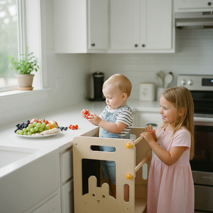 A toddler boy standing in the Hicooo learning tower holding cherry tomatoes while his older sister stands beside him at the kitchen counter with a plate of fresh fruit