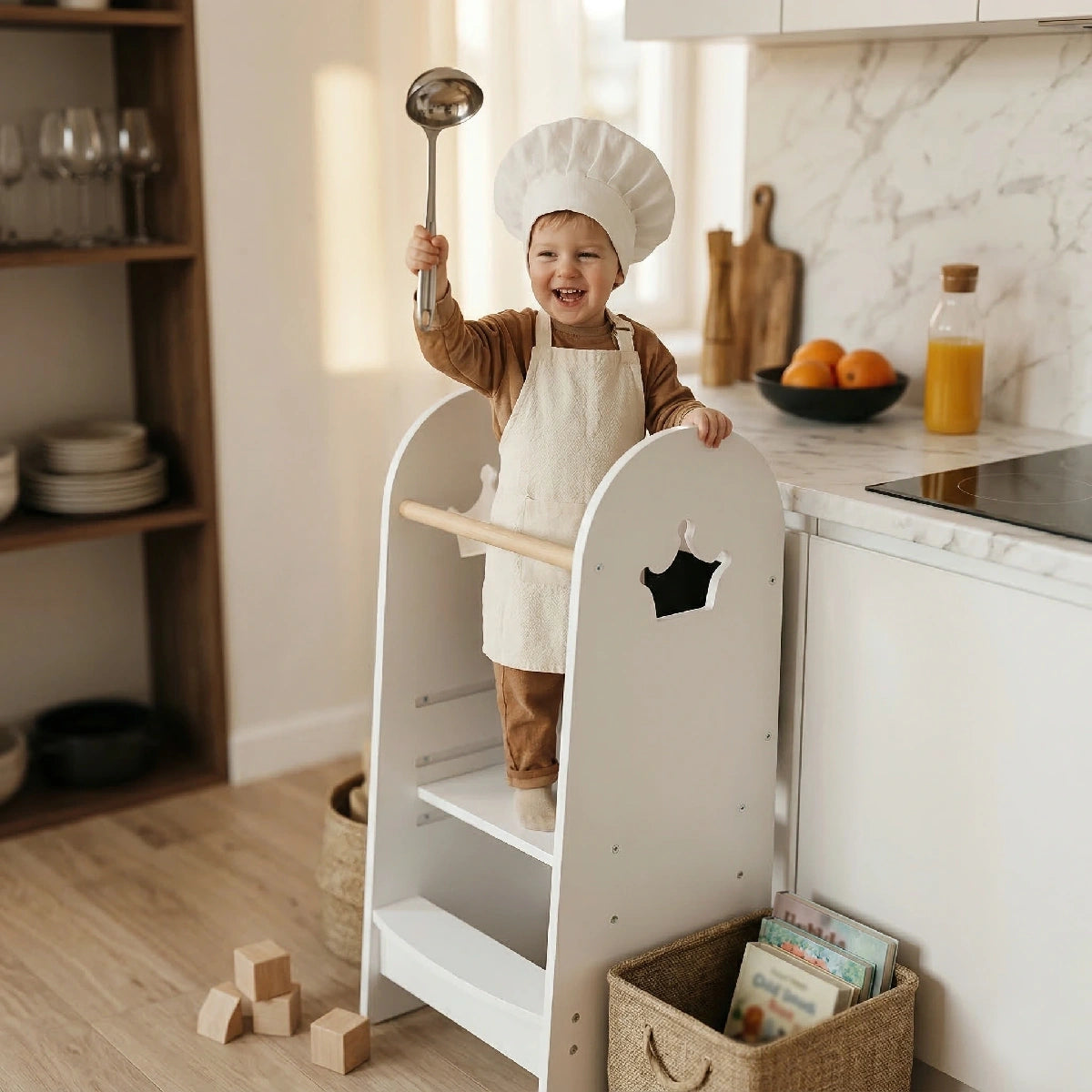 A laughing toddler boy wearing a chef's hat and apron stands safely in the Hicooo white wooden kitchen helper tower with crown cutouts, holding a ladle up high in a bright kitchen.