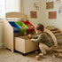 A toddler boy squatting on a woven rug in a sunlit playroom, putting a plush lion into the pulled-out rolling wooden toy box of a kids' storage organizer. A wooden climbing arch is naturally placed in the background, completing the cozy Montessori setup.