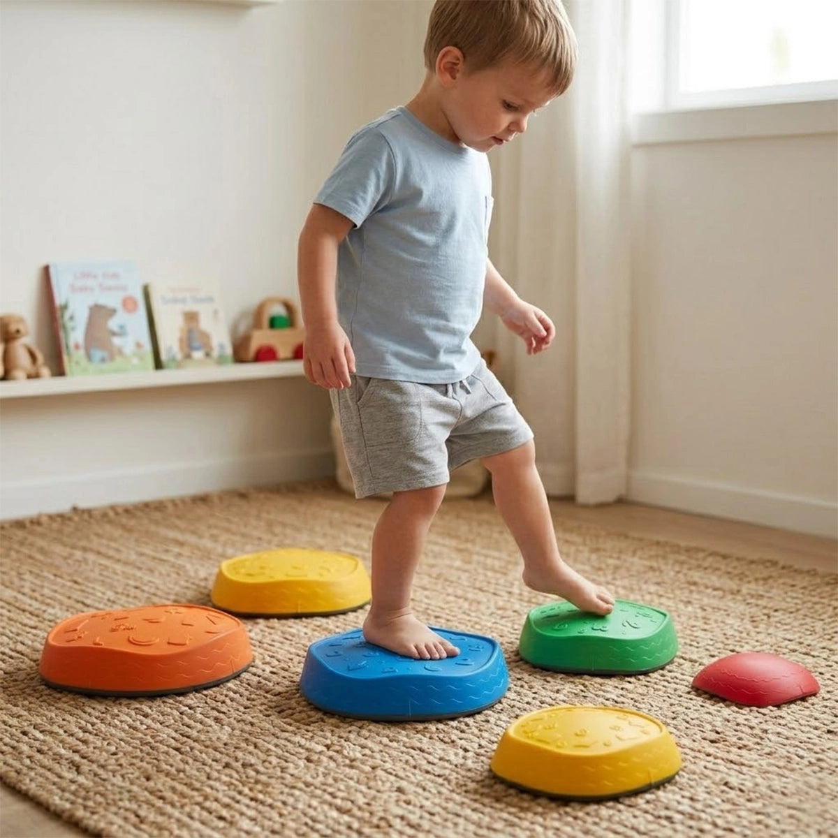 A barefoot toddler boy in a light blue t-shirt and grey shorts is carefully stepping from a large blue balance stone onto a green textured stone. He is playing on a woven rug in a brightly lit playroom. Several other colorful balance stepping stones in orange, yellow, and red are scattered around him. The background shows a low white shelf with children's books and wooden toys, illustrating a fun indoor activity that promotes gross motor skills and coordination.