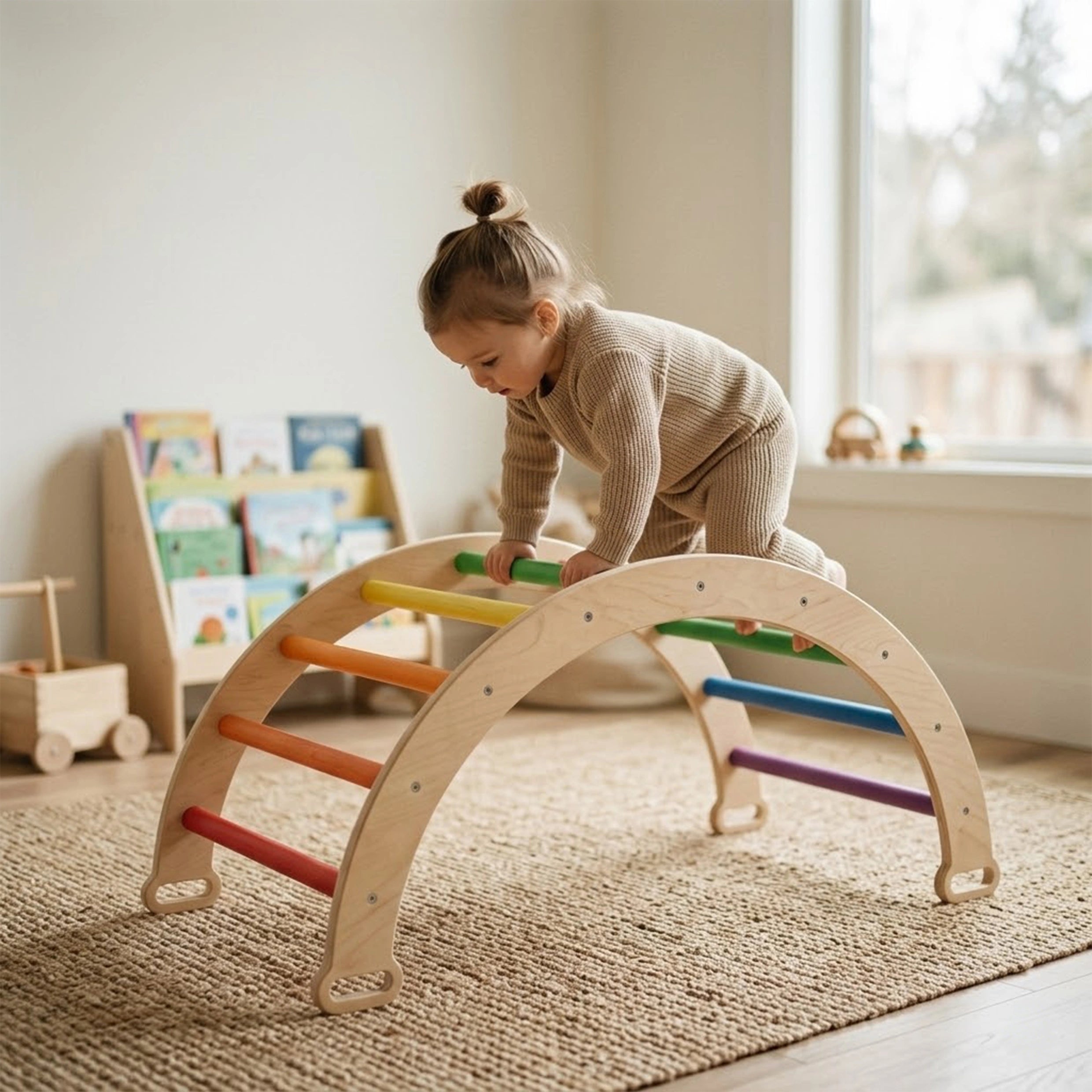 A toddler girl wearing a beige shirt and polka-dot pants actively climbing the wooden ramp of a Montessori Pikler triangle set. The sturdy indoor climbing frame features beautiful pastel rainbow-colored rungs and is placed on a textured rug in a bright, modern playroom.