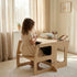A young girl with curly hair sits at the Hicooo convertible wooden desk in a sunlit room, drawing with crayons. Books and art supplies are neatly arranged on the desk's flat surface.