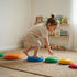 A young toddler girl with her hair in a bun, wearing beige play clothes, is balancing barefoot on a blue textured stepping stone. She is leaning forward, reaching out to touch a green stone placed on a woven rug. Colorful orange, yellow, and red stepping stones are also visible in the bright playroom, which features wooden toy shelves in the background. The image highlights active sensory play and coordination.