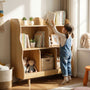 A toddler girl reaching up to pick a book from the top shelf of a wooden kids' bookshelf with wavy edges. Demonstrates the accessible height of this Montessori toy organizer.