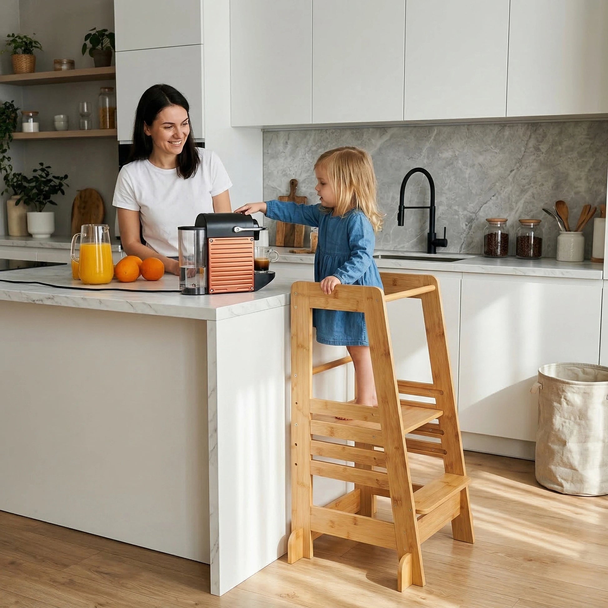 A toddler girl in a blue dress stands safely in the Hicooo natural bamboo learning tower at a kitchen island, reaching for a coffee maker while her smiling mother watches her closely.