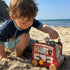 A young boy playing with a red wooden activity bus toy on a sandy beach during summer.