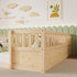 A light natural wood twin bed frame with a built-in guard rail, styled in a nursery setting with a green scalloped wall, wooden cloud decals, and a framed art print.