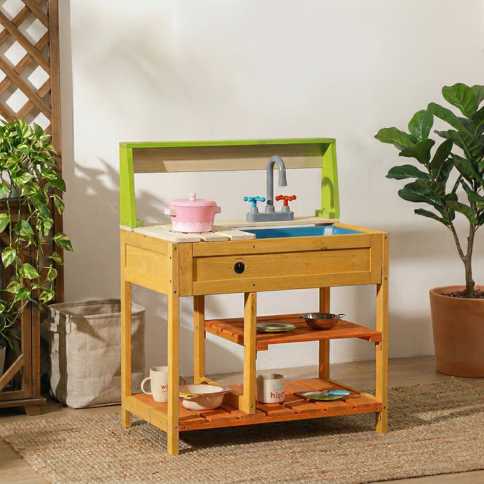A lifestyle photograph of a kids wooden mud kitchen set placed indoors on a woven rug. The play kitchen features a green top rack, a grey faucet with red and blue handles, a blue sink basin, and a stovetop. It is styled with various toy pots, plates, and mugs on its slatted shelves, next to a potted plant and a laundry basket.