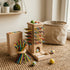 A lifestyle photograph capturing the completed and fully assembled wooden rainbow rod tower toy standing on a textured woven jute rug. The colourful stacked rings create a vibrant pyramid shape, and the wooden rods are neatly arranged within the base block. The softly blurred nursery playroom background includes a shelf with books and a small potted plant.