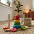 A classic wooden Tower of Hanoi puzzle featuring vibrant, colorful stacking rings on the center peg. The toy rests on a textured woven rug inside a cozy, modern nursery, with a blurred background showing a potted plant and natural sunlight.